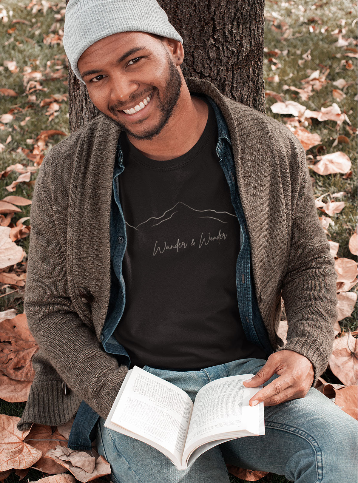 Person wearing unisex mountain graphic t-shirt, sitting under a tree, happily reading book in grey t-shirt 