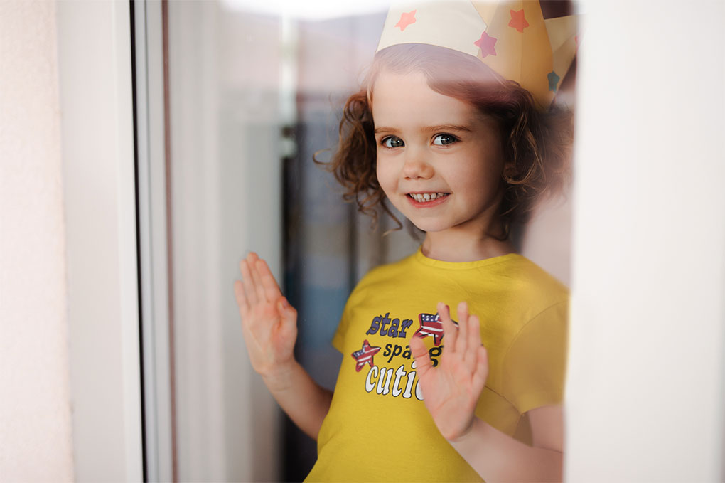 Toddler looking out the  storm door wearing a paper crown and smiling- wearing a bright yellow "star spangled cutie" tee