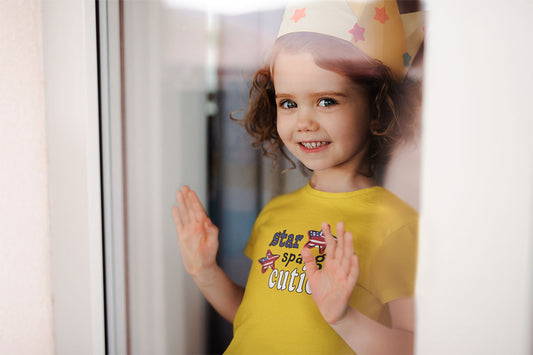 Toddler looking out the  storm door wearing a paper crown and smiling- wearing a bright yellow "star spangled cutie" tee