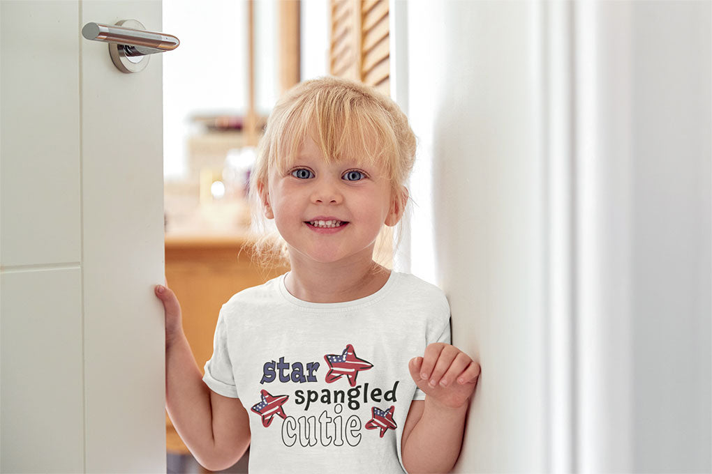 Toddler opening the front door with a sweet smile - wearing a white "star spangled cutie" tee