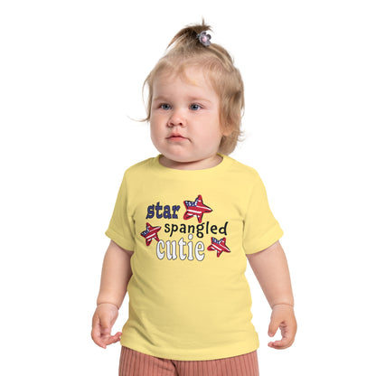 Baby wearing a light yellow t-shrit with typography and patriotic stars, t-shirt says "star spangled cutie"